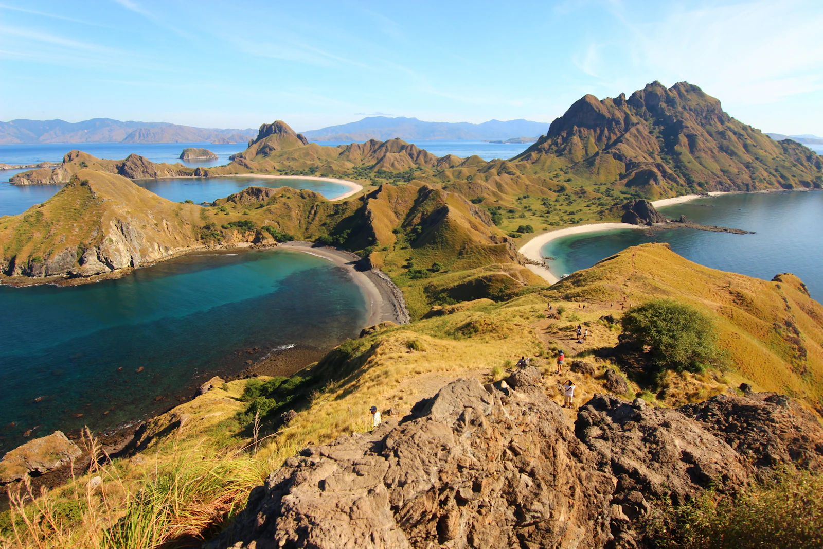 Padar Island's hills and bays seen from the rocky viewpoint at the top of the hiking trail, with hikers on the path below
