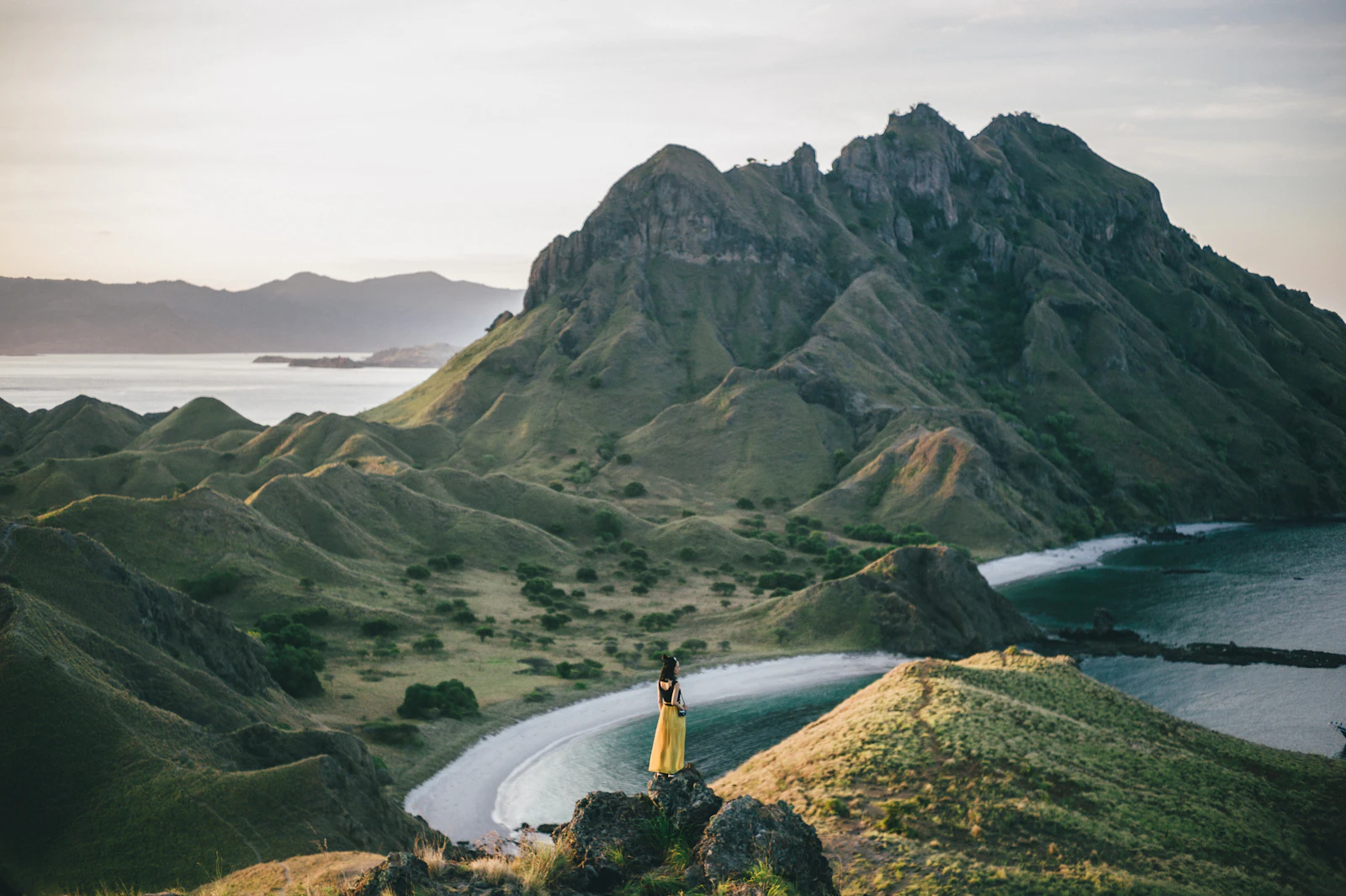 A visitor standing on a rocky outcrop on Padar Island's ridge, looking out over a curved bay and the island's peaks