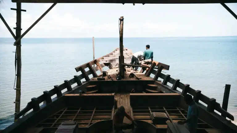 The unfinished frame of a wooden phinisi under construction on the shore
